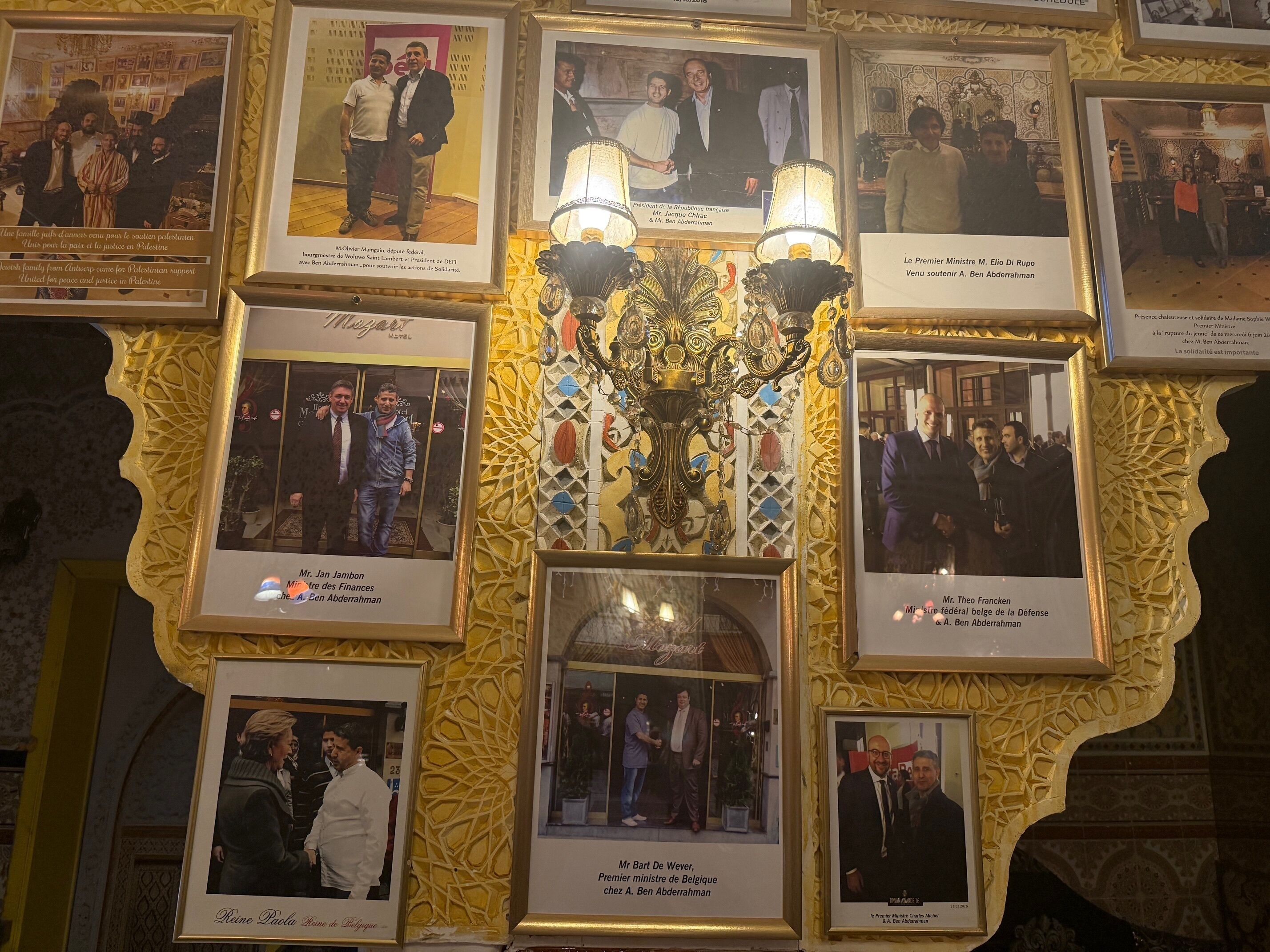 A wall with framed photographs and text describing various individuals and their roles, surrounded by ornate golden frames and a chandelier.
