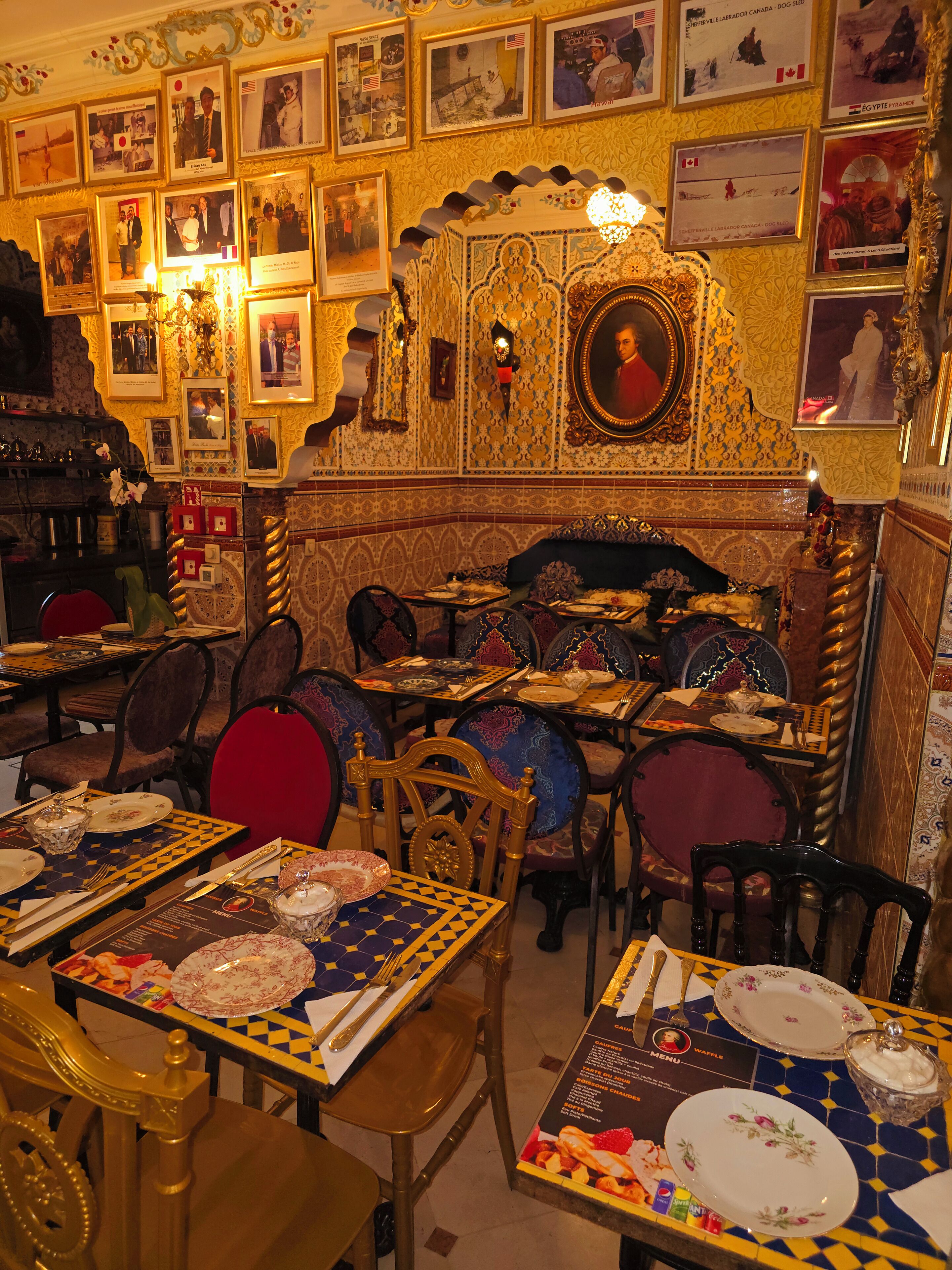 Un restaurante con una decoración elaborada, que incluye cuadros enmarcados en las paredes y un piso de baldosas con diseños. Las mesas están dispuestas con platos, cubiertos y menús.