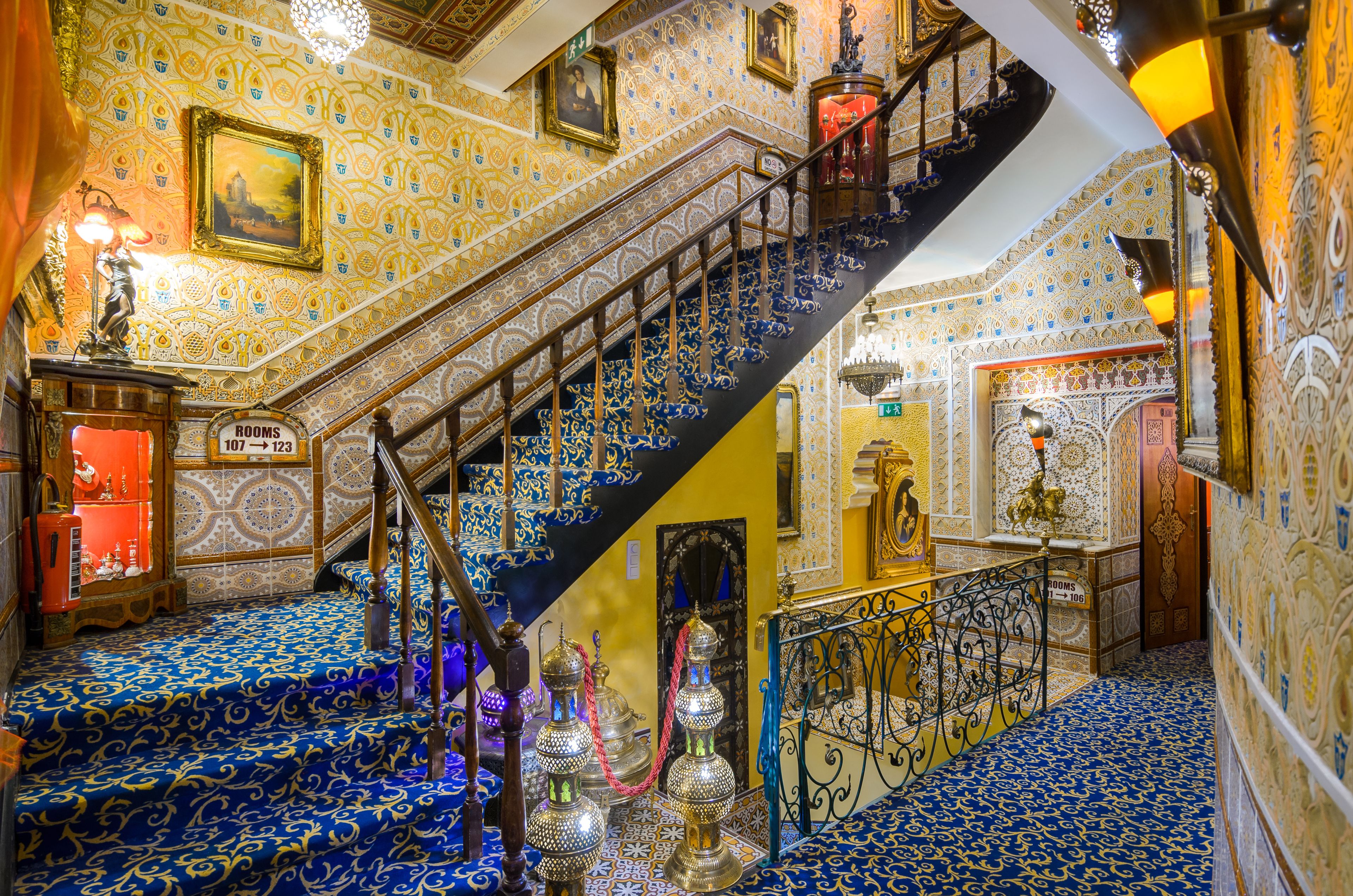 A staircase with ornate blue and gold patterns, a sign indicating rooms 107 to 123, and a chandelier.