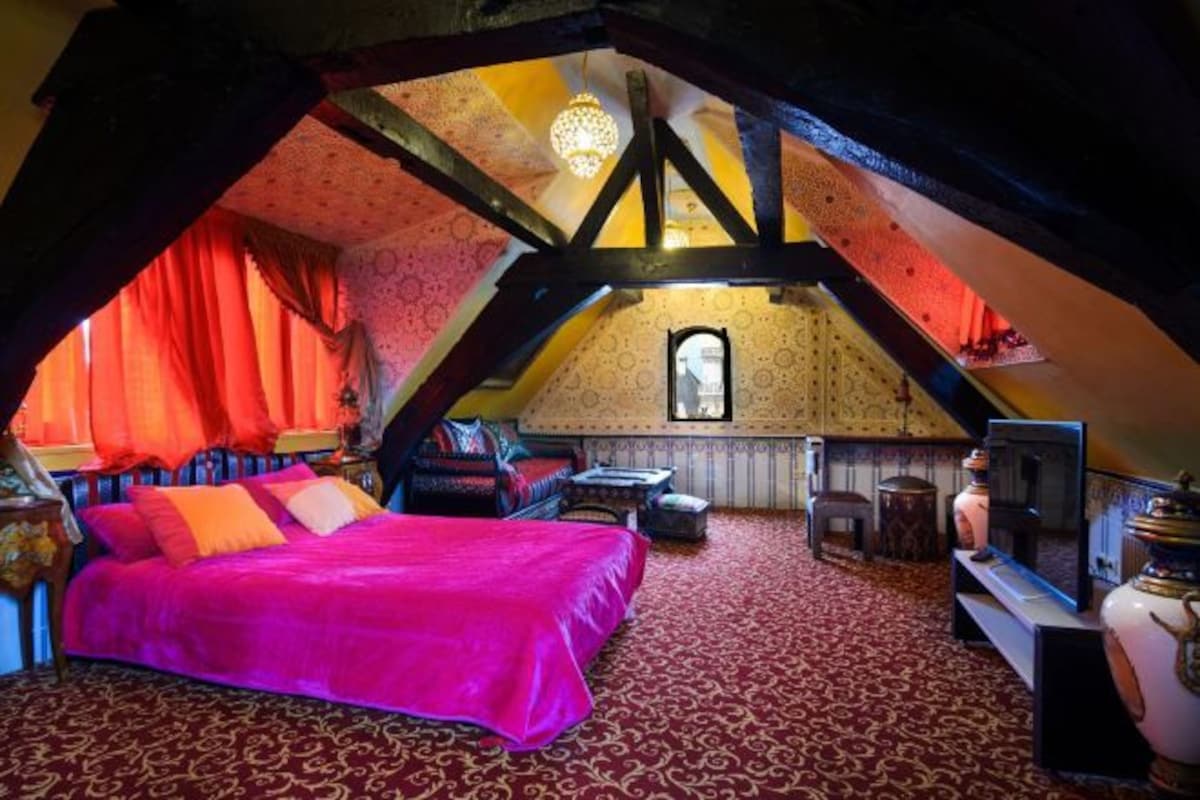 An attic bedroom with a bed, a small table, a chair, a TV, and a decorative vase.