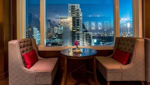 A room with a large window offering a city view, two upholstered chairs, a small round table, and a vase with flowers. - Hilton Bangkok Grande Asoke (Bangkok)