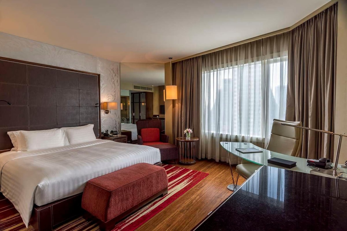 Une chambre d’hôtel avec un grand lit, un bureau, une chaise, un pouf rouge et une vue sur la ville par la fenêtre.