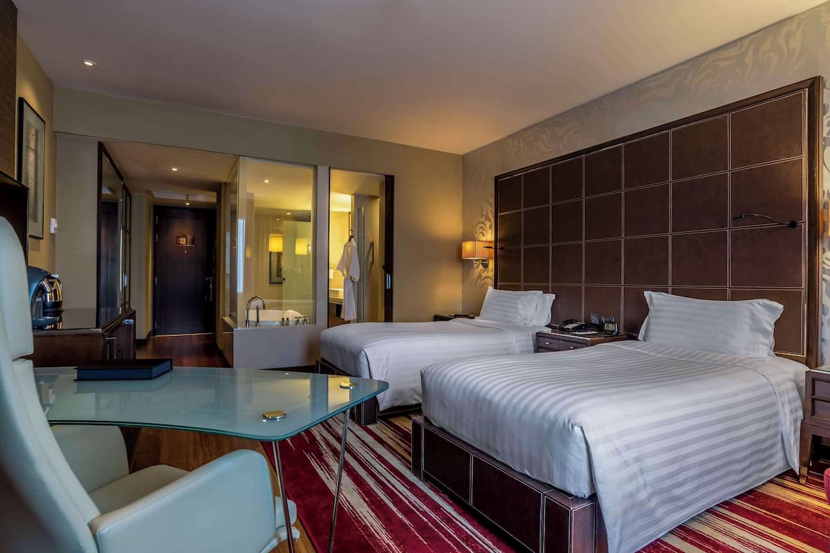 Hotelkamer met twee bedden, een glazen tafel, een stoel, een badkamer met bad en een grote spiegel.