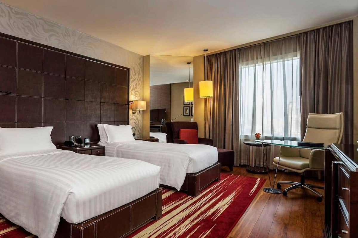 Een hotelkamer met twee bedden, een bureau, een stoel en een groot raam met gordijnen.