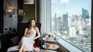A woman sitting by a window with a cityscape view, a tiered tea service, and a lamp on the wall. - Hilton Bangkok Grande Asoke (Bangkok)