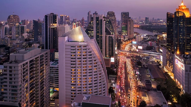 A high-rise hotel with a distinctive curved roof, located in a bustling cityscape with a busy road and other tall buildings in the background. - Hilton Bangkok Grande Asoke (Bangkok)