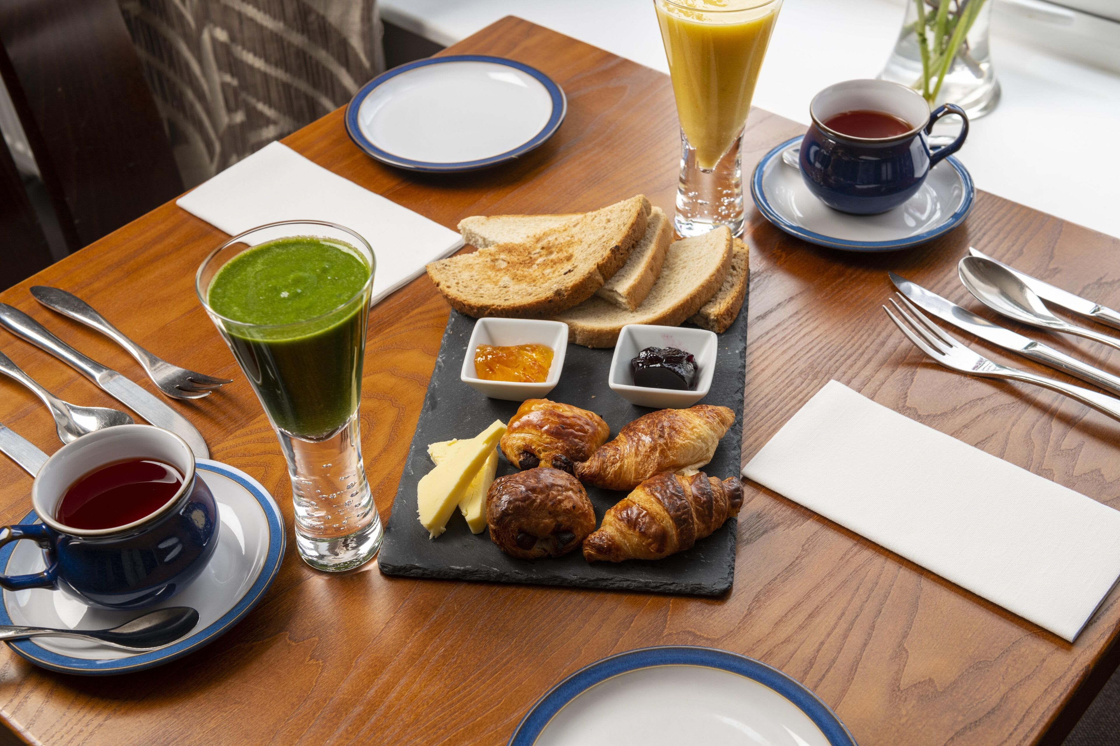 A breakfast setting with toast, croissants, jam, butter, and a green smoothie on a slate board.