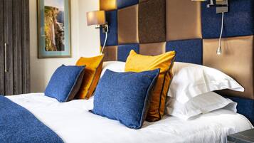 Habitación de hotel con una cama grande, almohadas azules y amarillas, cabecera con estampado a cuadros y un cuadro en la pared.