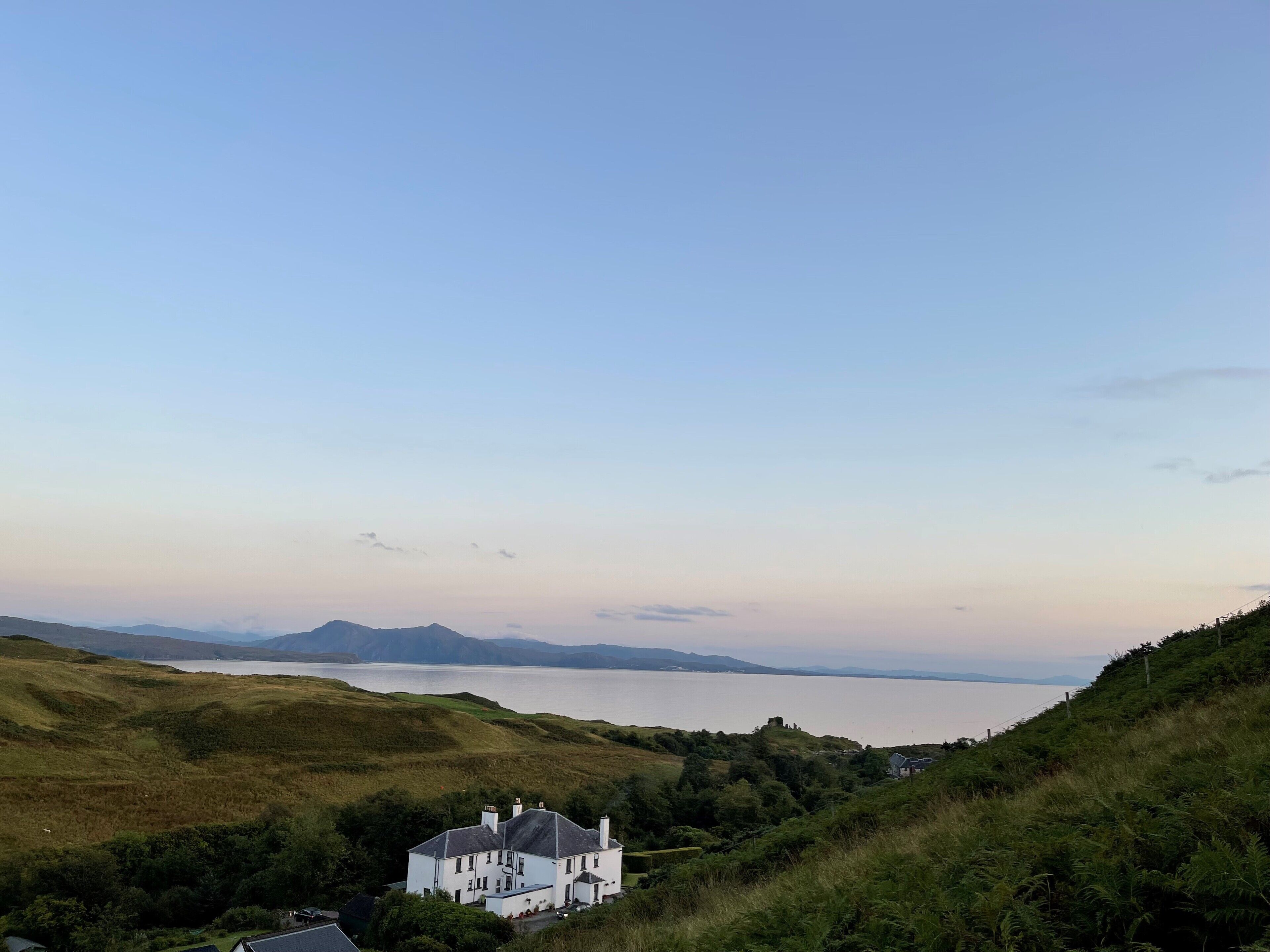 A white house with a steep roof, surrounded by lush green hills and a body of water in the distance.