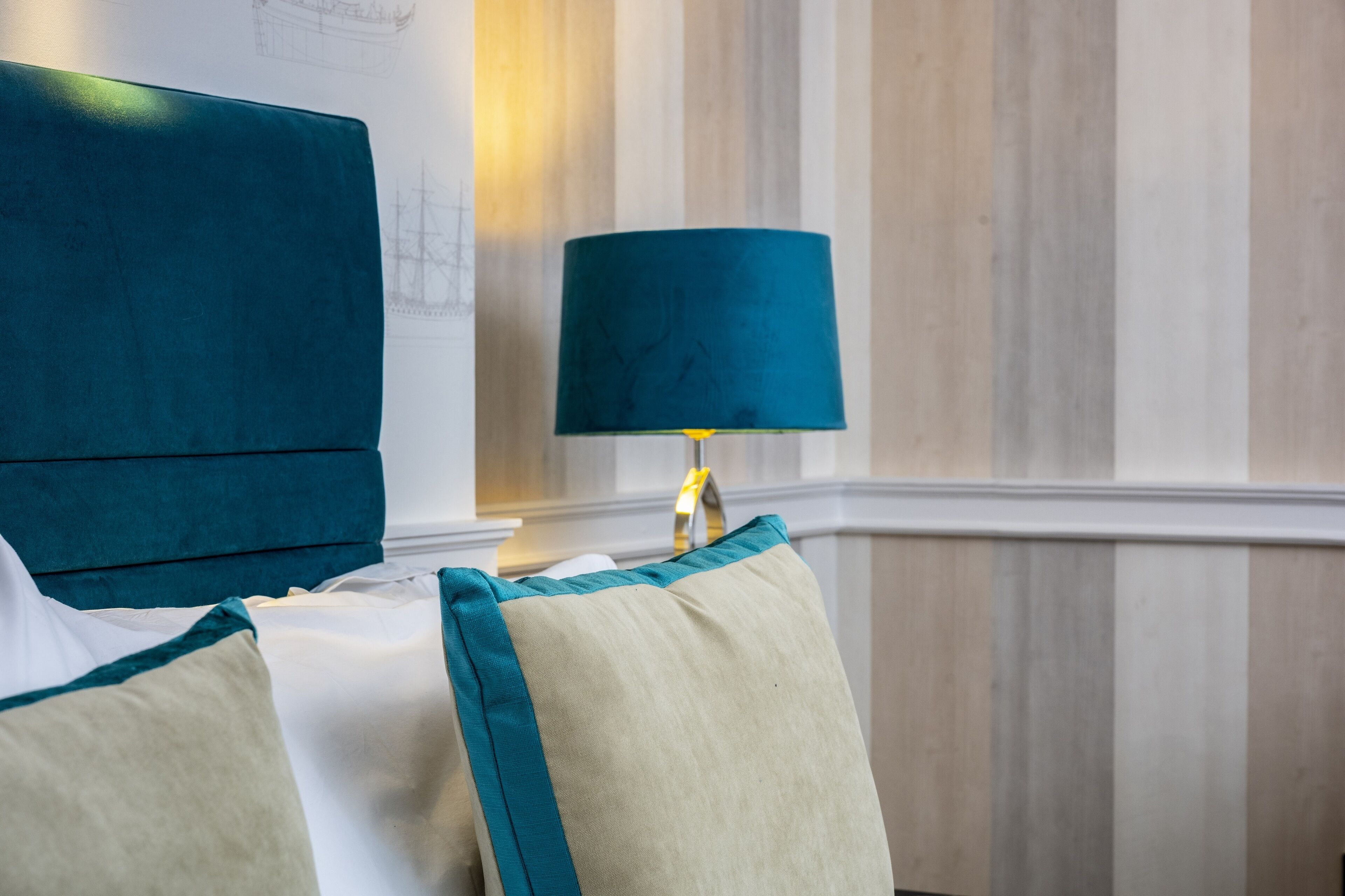 Habitación de hotel con una cama, un cabecero tapizado en color turquesa, un cojín color crema y una pantalla de lámpara turquesa sobre una mesita de noche.
