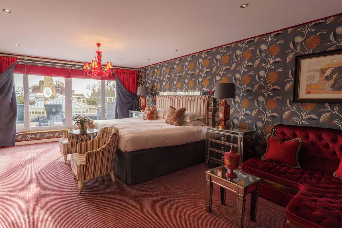 Quarto com uma cama grande, um sofá vermelho, uma mesa pequena e uma cadeira. Há uma janela com cortinas e vista para edifícios.