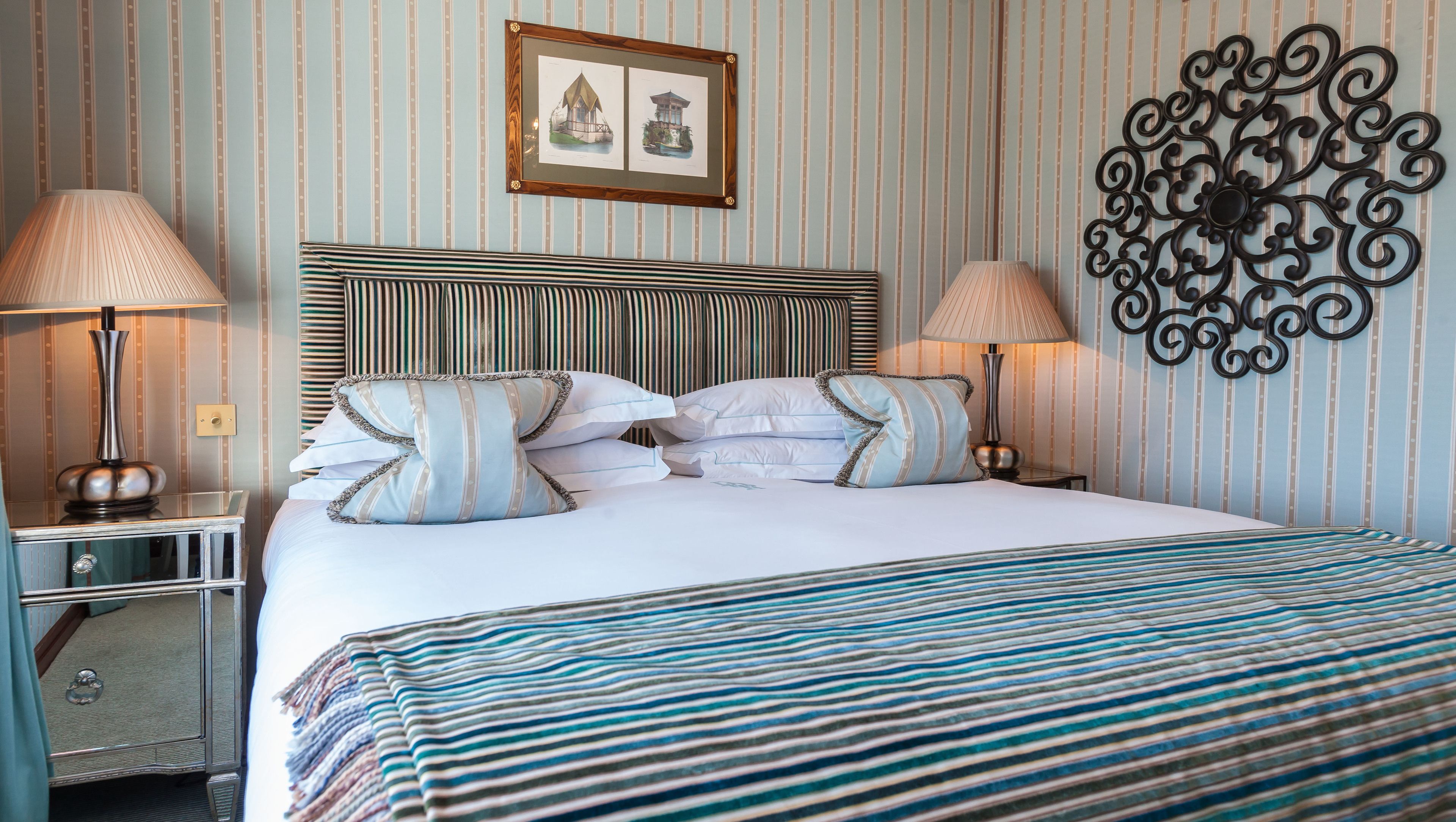 A hotel room with a striped wallpaper, a bed with striped bedding, two bedside lamps, a decorative wall piece, and a framed picture on the wall.