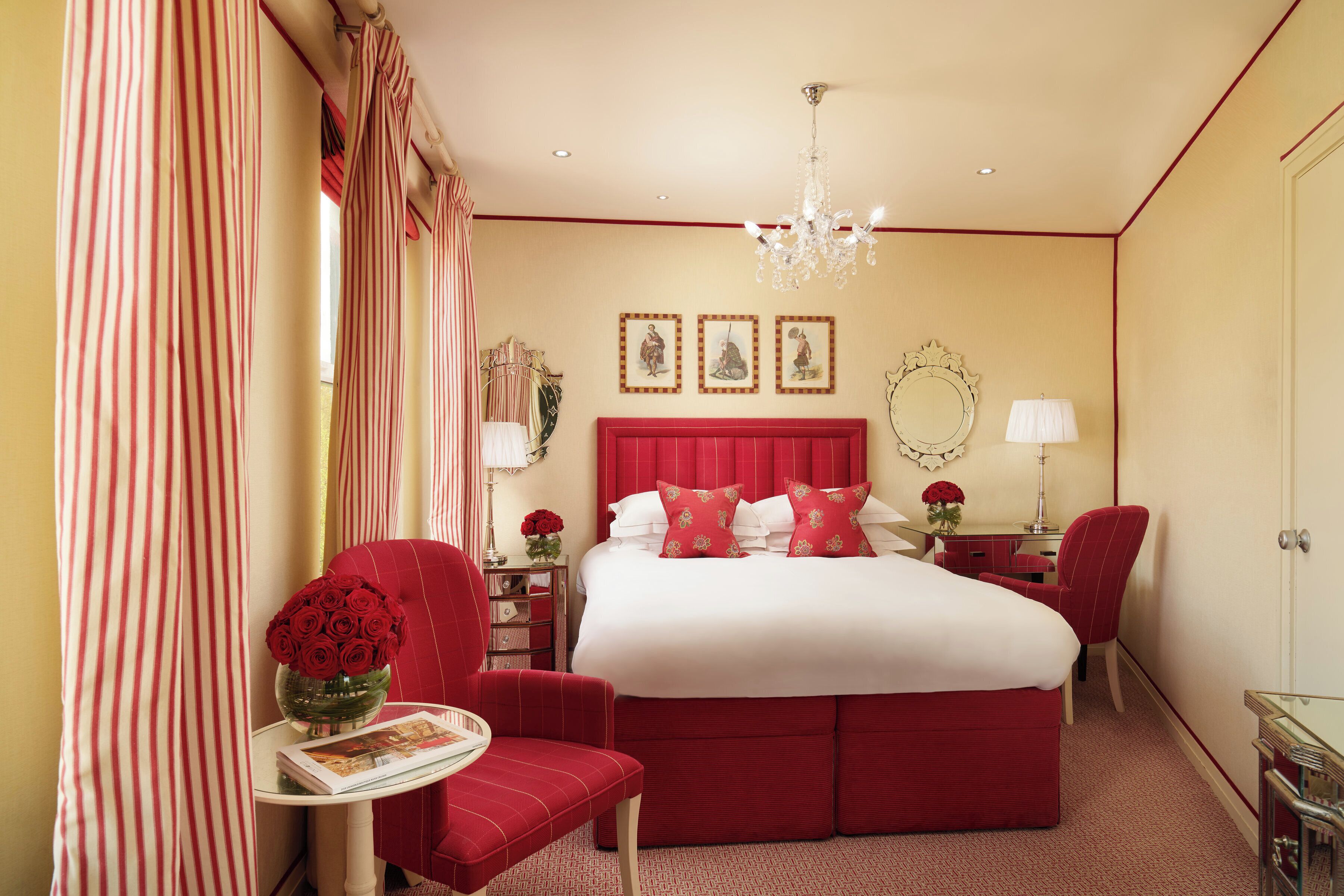 A hotel room with a large bed, a red upholstered chair, a small table with a vase of red flowers, and a desk with a lamp.