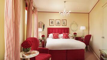 A hotel room with a large bed, a red upholstered chair, a small table with a vase of red flowers, and a desk with a lamp.
