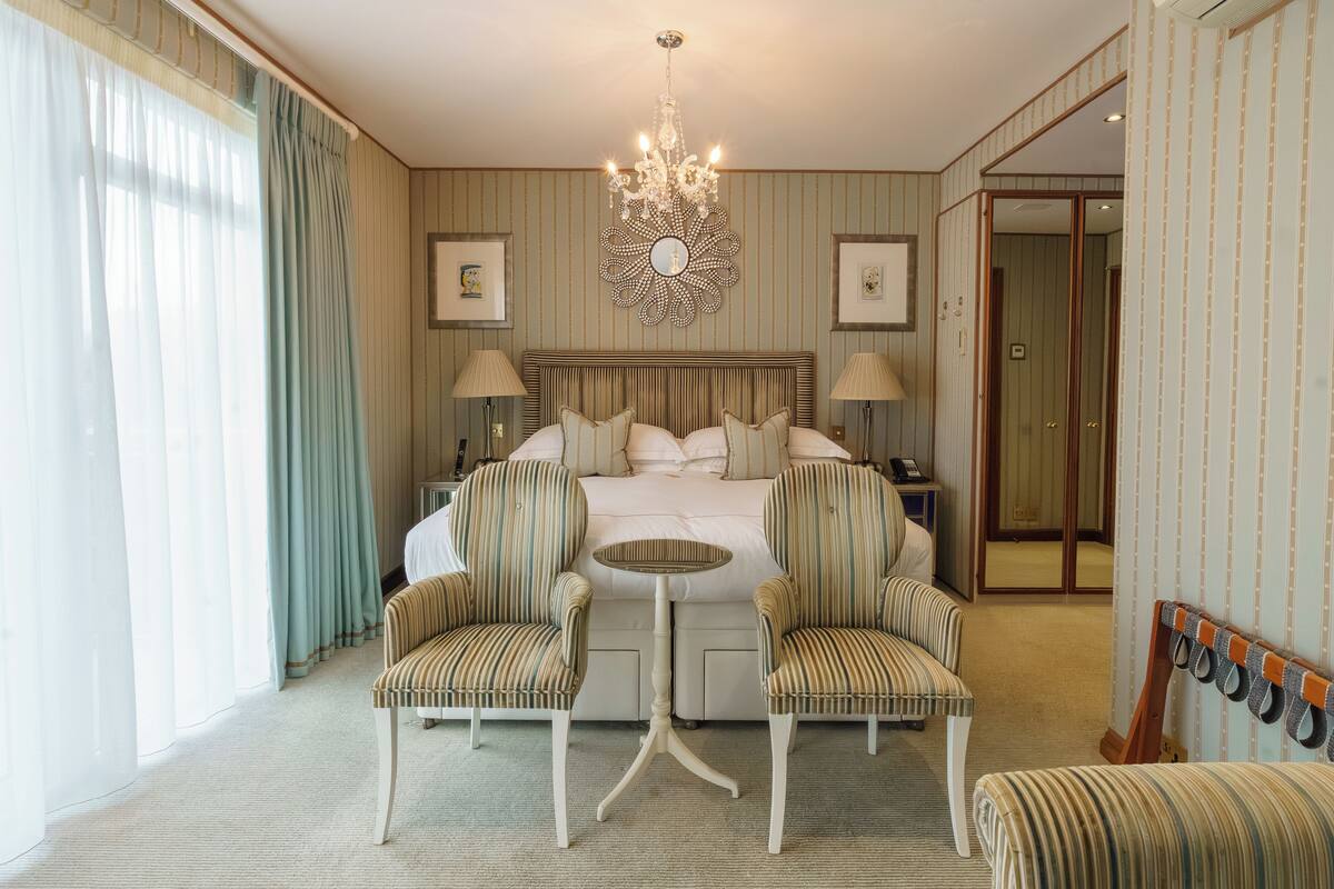 Quarto de hotel com uma cama grande, duas poltronas, uma mesa redonda pequena e um candelabro.
