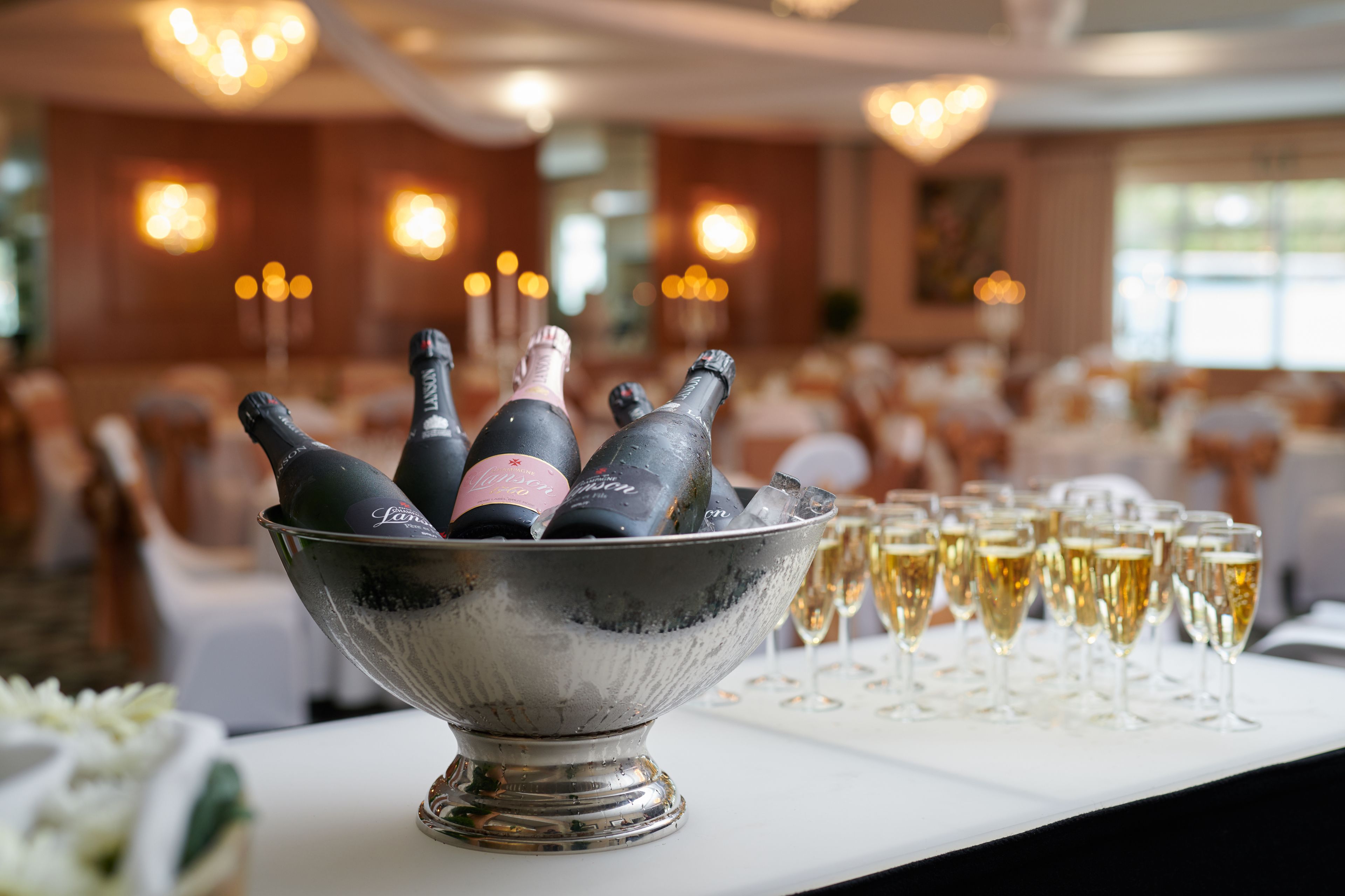 A table with a silver bowl containing champagne bottles and glasses, set in a banquet hall with chandeliers and white tables.