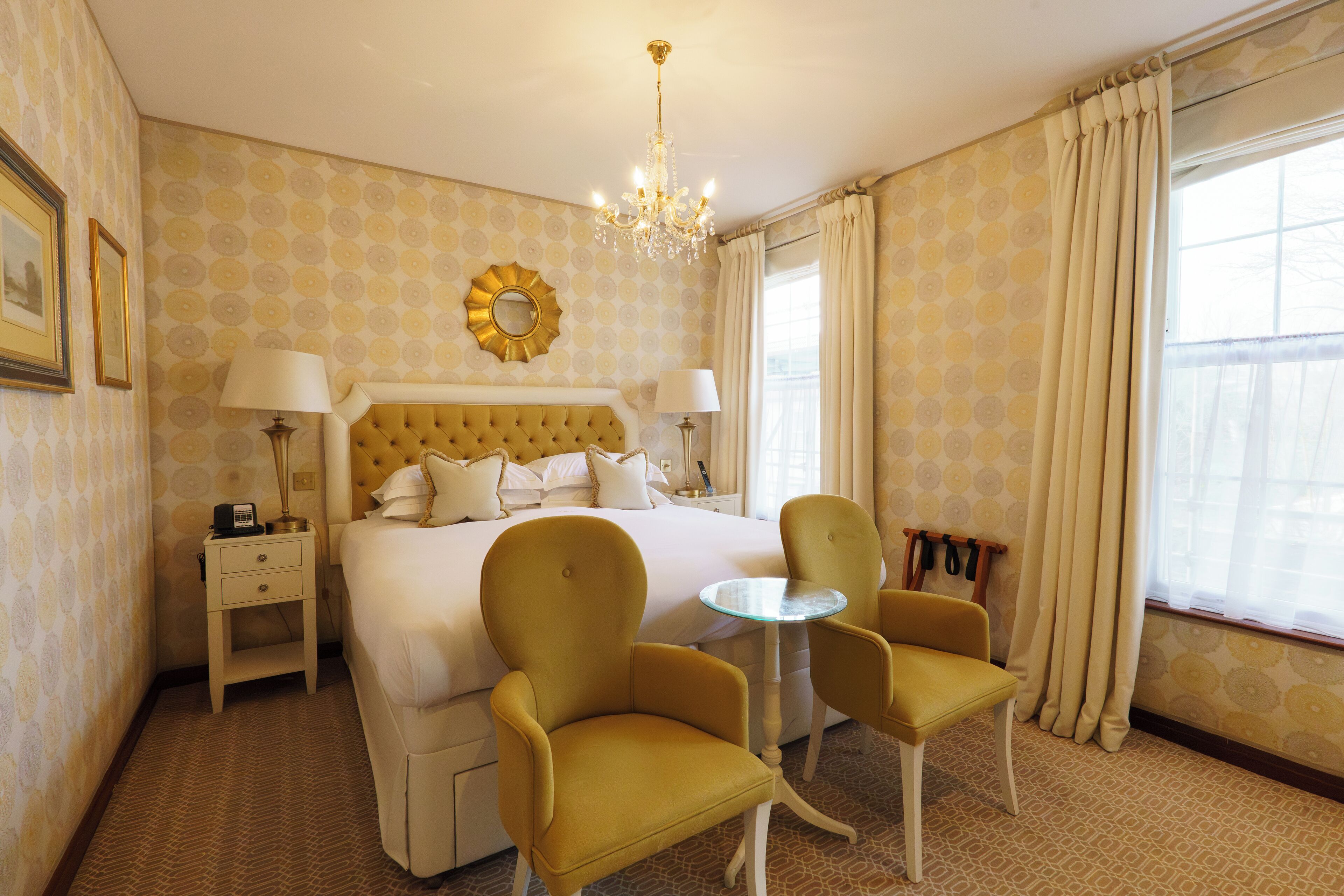Ein Hotelzimmer mit einem großen Bett, zwei Sesseln, einem kleinen runden Tisch, einer Chandelieber, Nachttischen mit Lampen und einem Spiegel an der Wand.