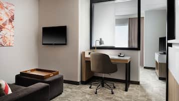 A modern hotel room with a flat-screen TV, a desk with a chair, a wooden side table, and a large mirror.