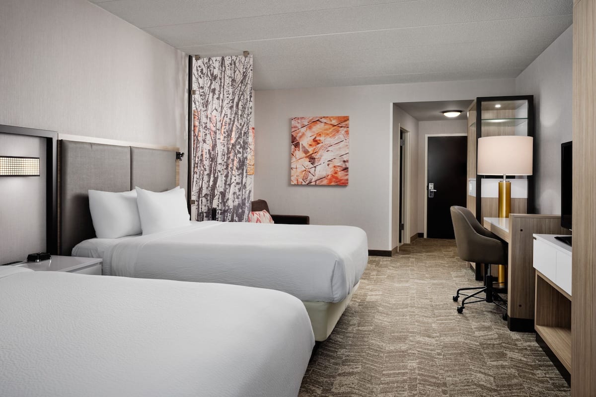 A hotel room with two beds, a desk, a chair, a lamp, and a painting on the wall.