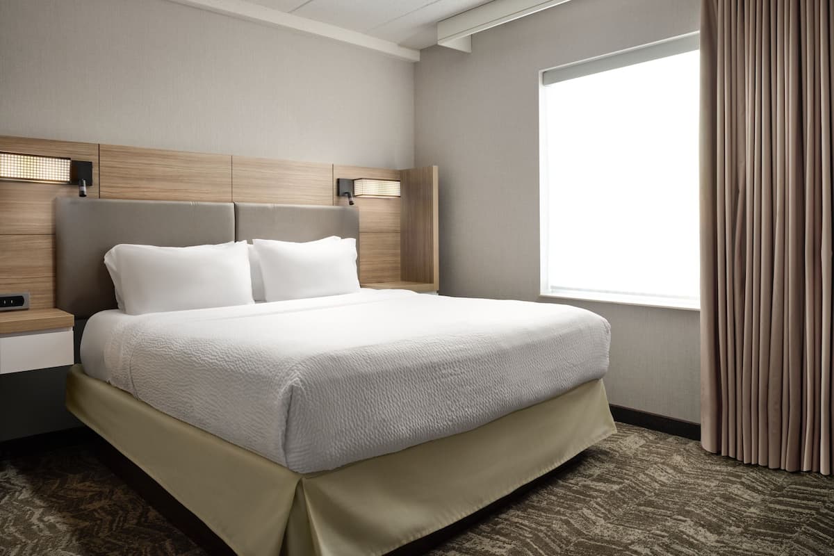 A hotel room with a large bed, a nightstand, a wall-mounted lamp, and a window with curtains.