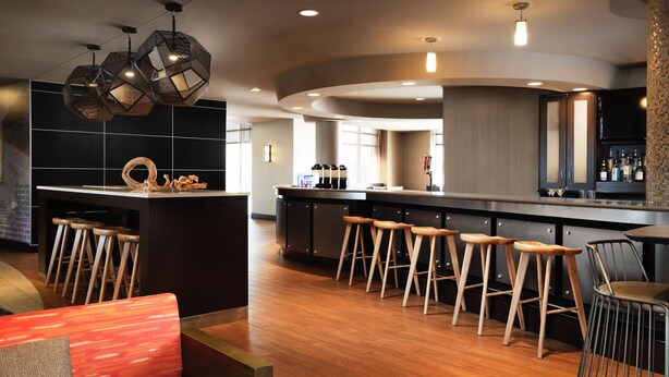 A modern bar area with wooden stools, a central bar counter, and a seating area with a red cushioned bench.