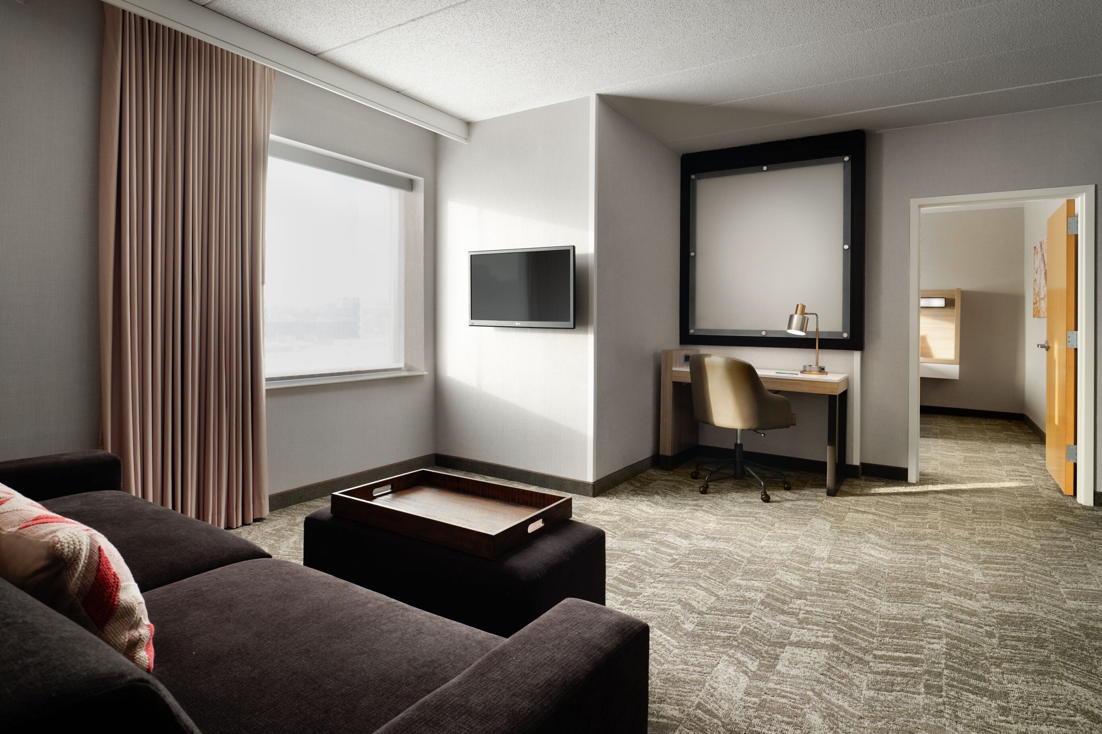 Habitación de hotel con sofá, escritorio, televisor y un ventanal con cortinas.