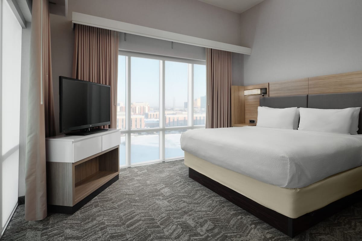 A modern hotel room with a large bed, a flat-screen TV on a wooden console, a bedside table, and a view of the city through floor-to-ceiling windows.