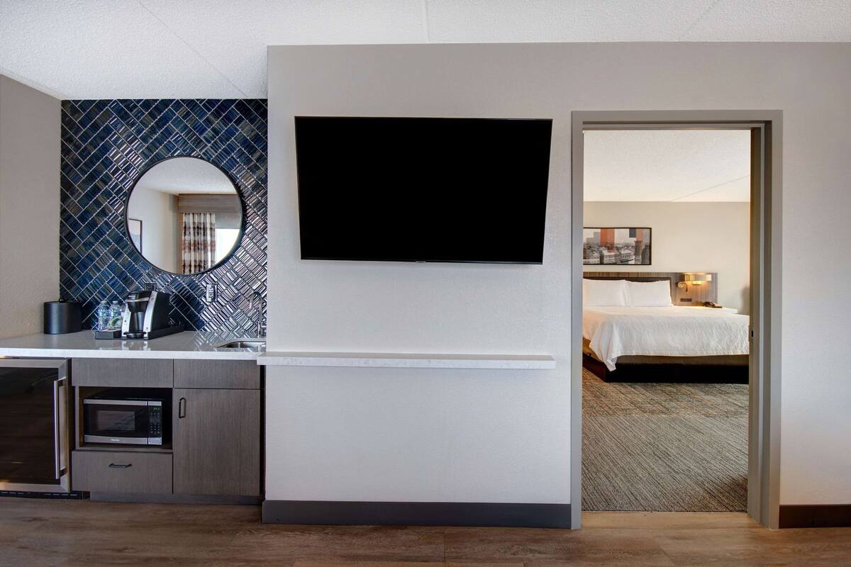A hotel room with a flat-screen TV mounted on the wall, a bed with white bedding, and a view of the cityscape through the window.