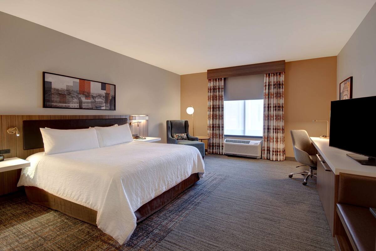 A hotel room with a large bed, a desk with a computer, a TV, and a window with curtains.
