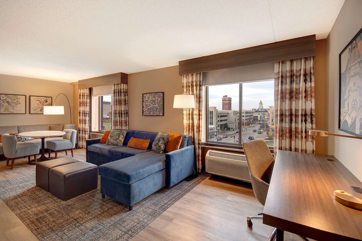 A modern hotel room with a blue sofa, a dining table, and a large window offering a city view.