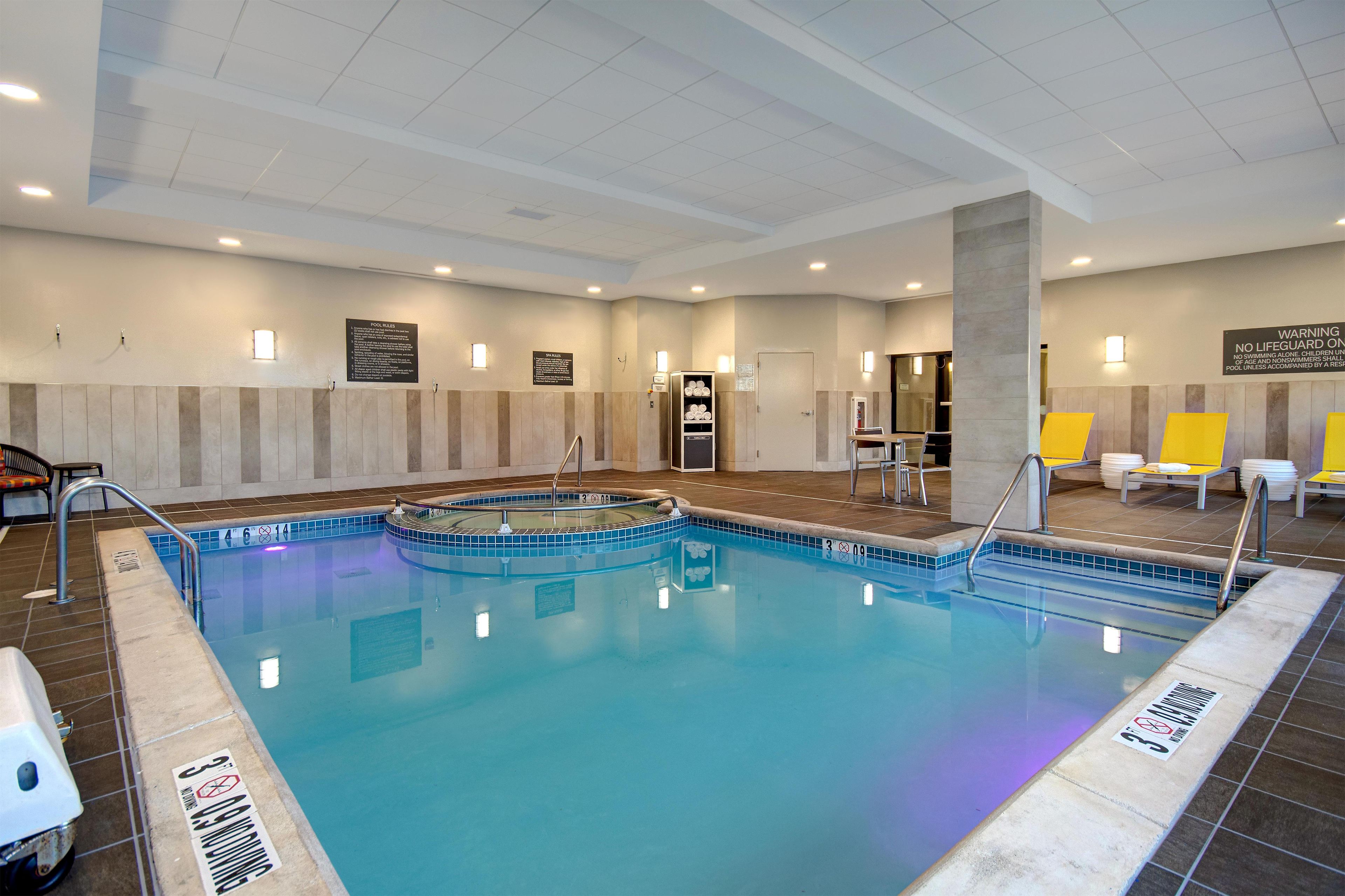 An indoor swimming pool with clear water, tiled edges, and a pool ladder. There are lounge chairs and a sign indicating pool rules.