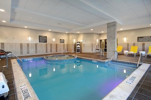 An indoor swimming pool with clear water, tiled edges, and a pool ladder. There are lounge chairs and a sign indicating pool rules.