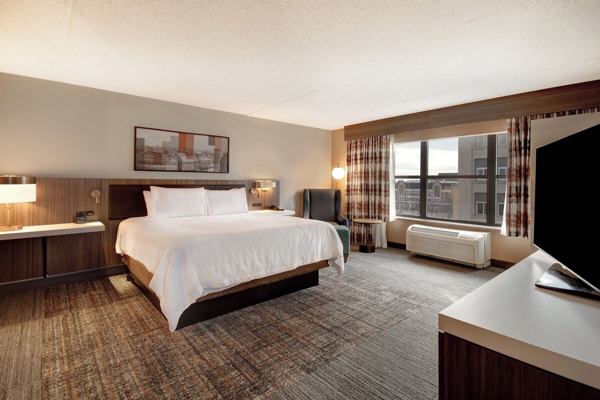 A hotel room with a large bed, a desk, a chair, a TV, a window with curtains, and a painting on the wall.