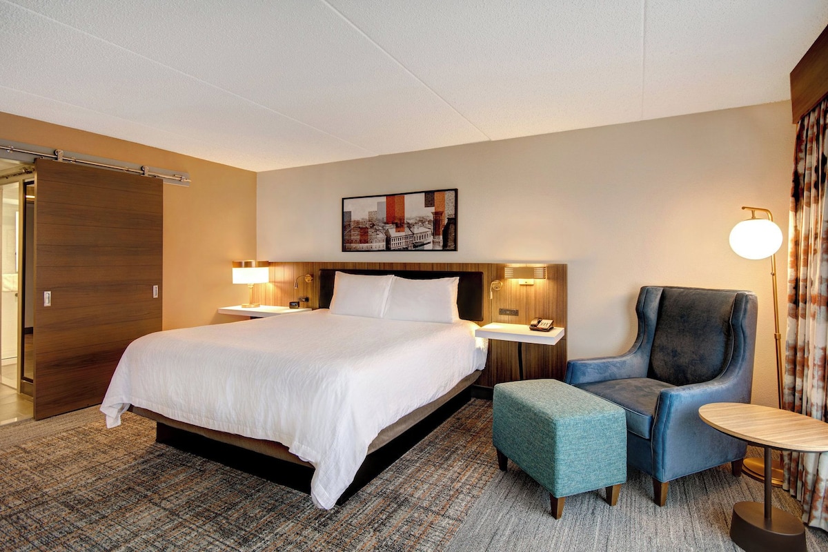 A hotel room with a large bed, bedside tables, a chair, a small table, a lamp, and a painting on the wall.