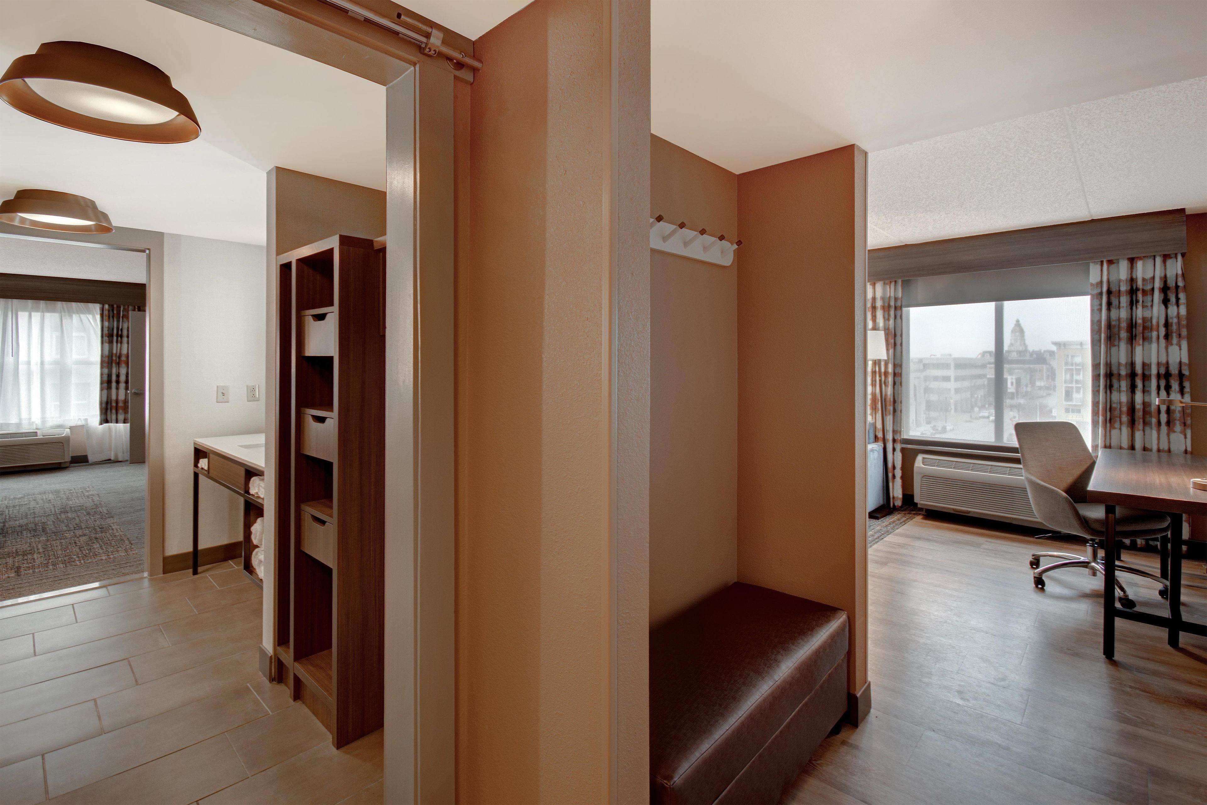 A hotel room with a wooden wardrobe, a desk, a chair, a bench, and a large window with a view.