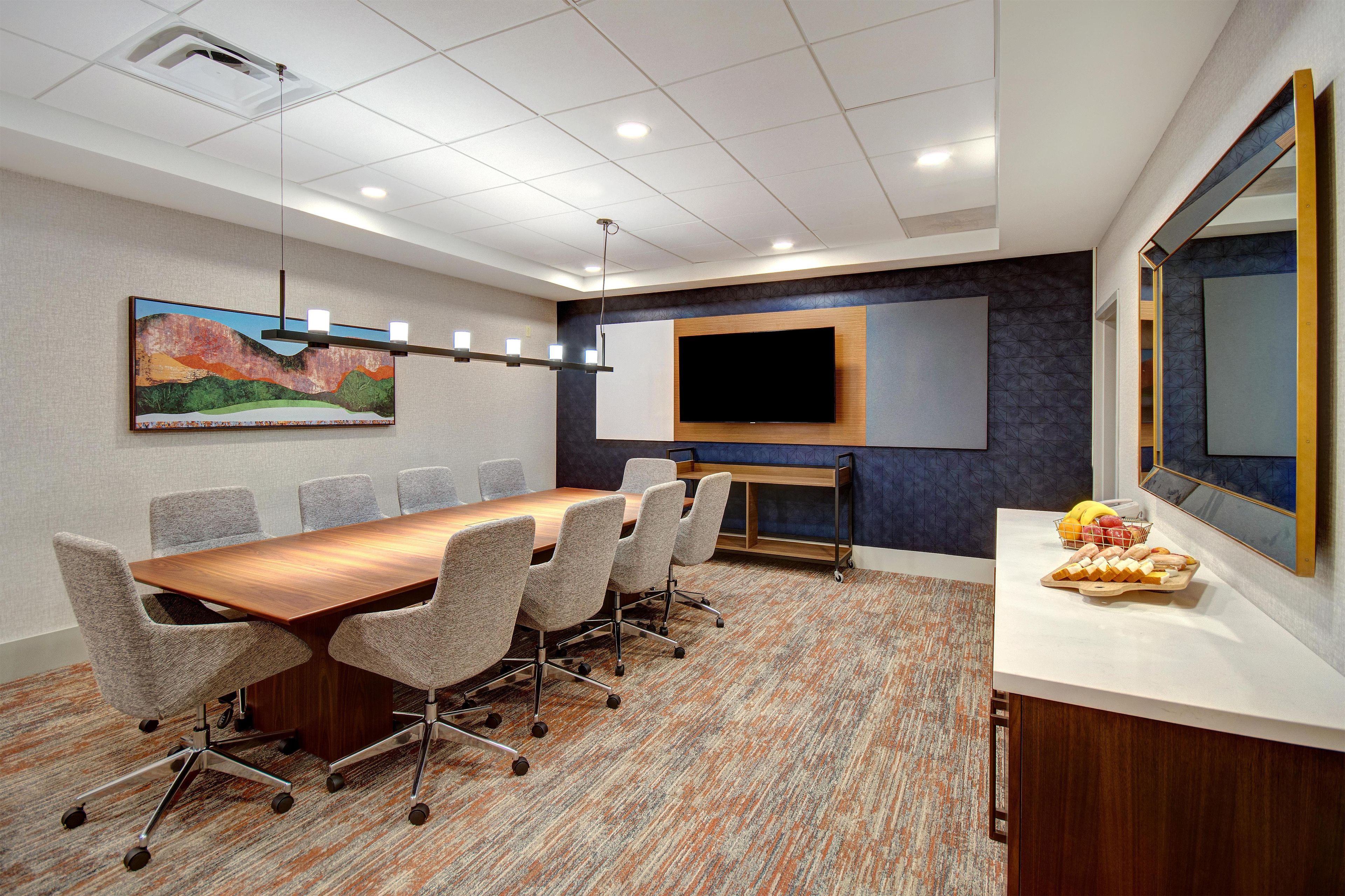 A conference room with a long table, six chairs, a flat-screen TV, a painting, and a fruit basket.