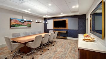 A conference room with a long table, six chairs, a flat-screen TV, a painting, and a fruit basket.