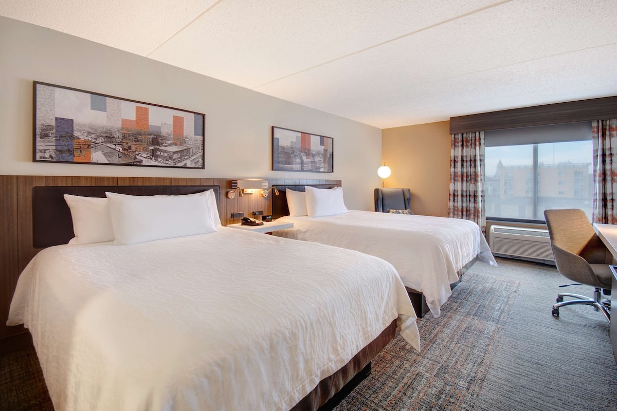 A hotel room with two beds, a desk, a chair, a window with curtains, and two wall-mounted artworks.