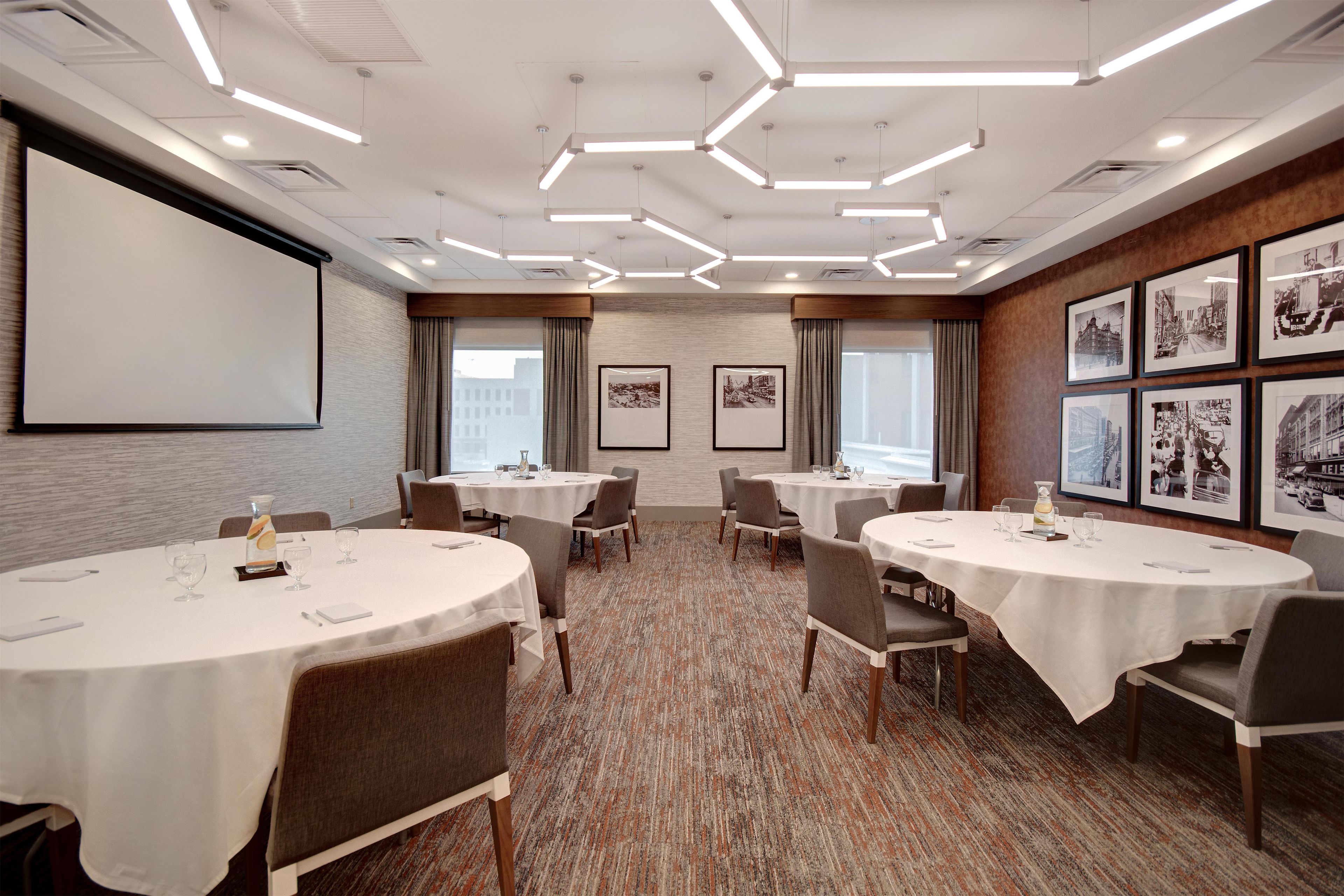A conference room with multiple tables, chairs, a projector screen, and framed pictures on the walls.