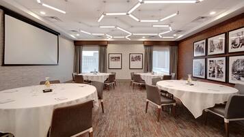 A conference room with multiple tables, chairs, a projector screen, and framed pictures on the walls.