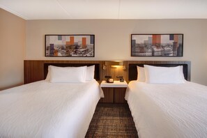 A hotel room with two beds, a nightstand with a lamp, and two framed cityscape pictures on the wall.