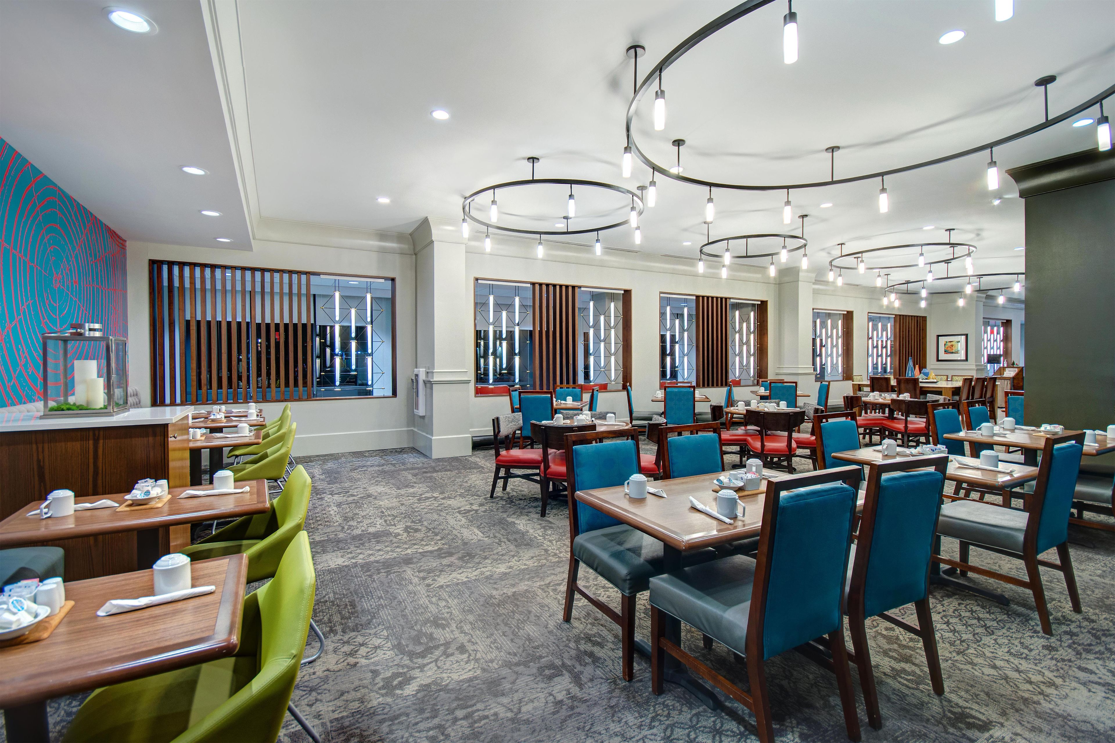 A hotel dining area with multiple tables and chairs, a patterned carpet, and a modern ceiling light fixture.