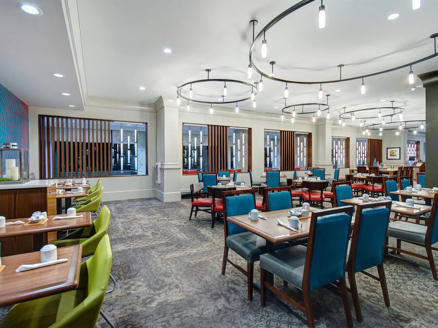 A hotel dining area with multiple tables and chairs, a patterned carpet, and a modern ceiling light fixture.