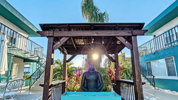 Une statue représentant une figure assise, placée sous une pergola en bois, avec un banc devant, le tout entouré de verdure et de bâtiments.