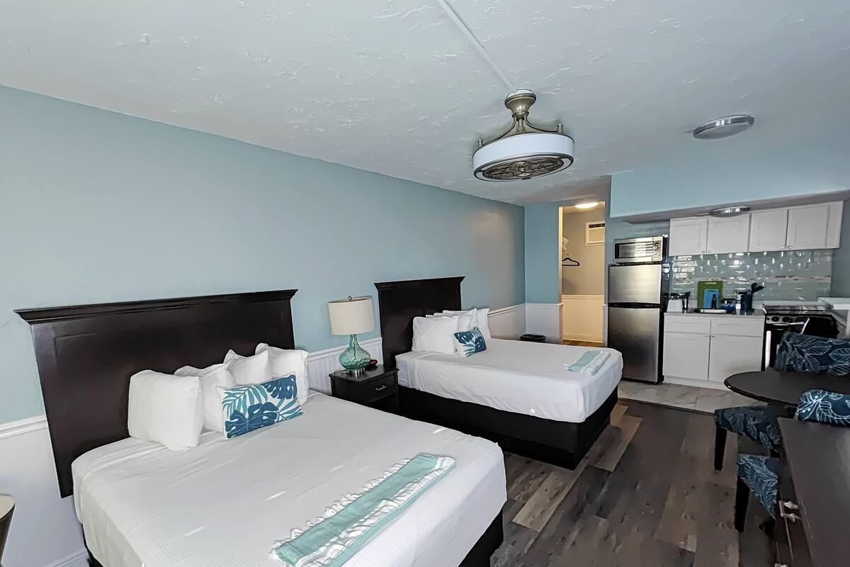 A hotel room with two beds, a small desk, a chair, a kitchenette, and a ceiling fan.