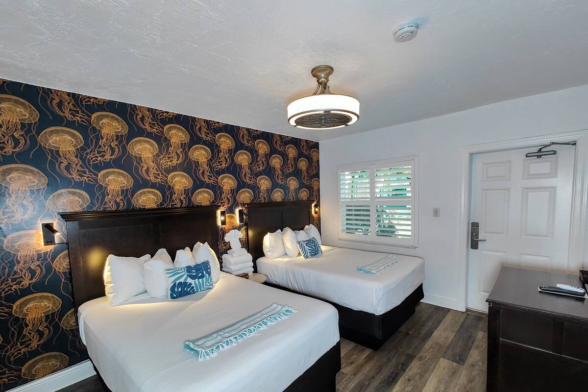 A hotel room with two beds, a ceiling fan, a window with blinds, and a wall with a jellyfish pattern.