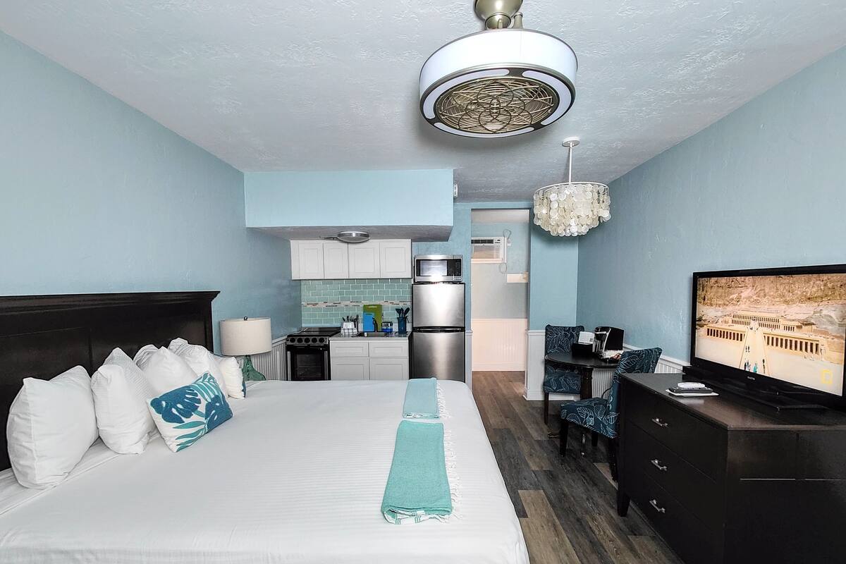A hotel room with a bed, a desk, a television, a kitchenette, and a ceiling fan.