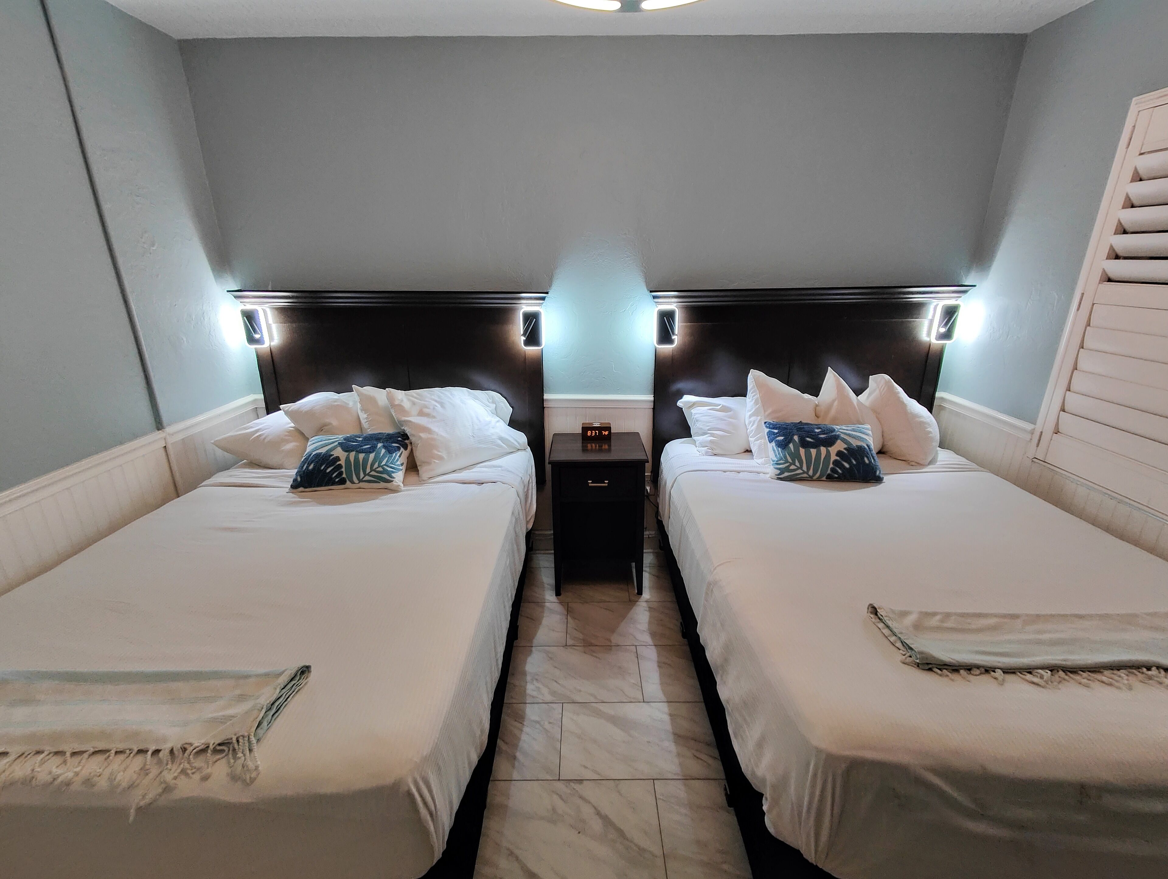 A mirrored room with two beds, each with white bedding and decorative pillows, a nightstand with a clock, and wall-mounted lighting.