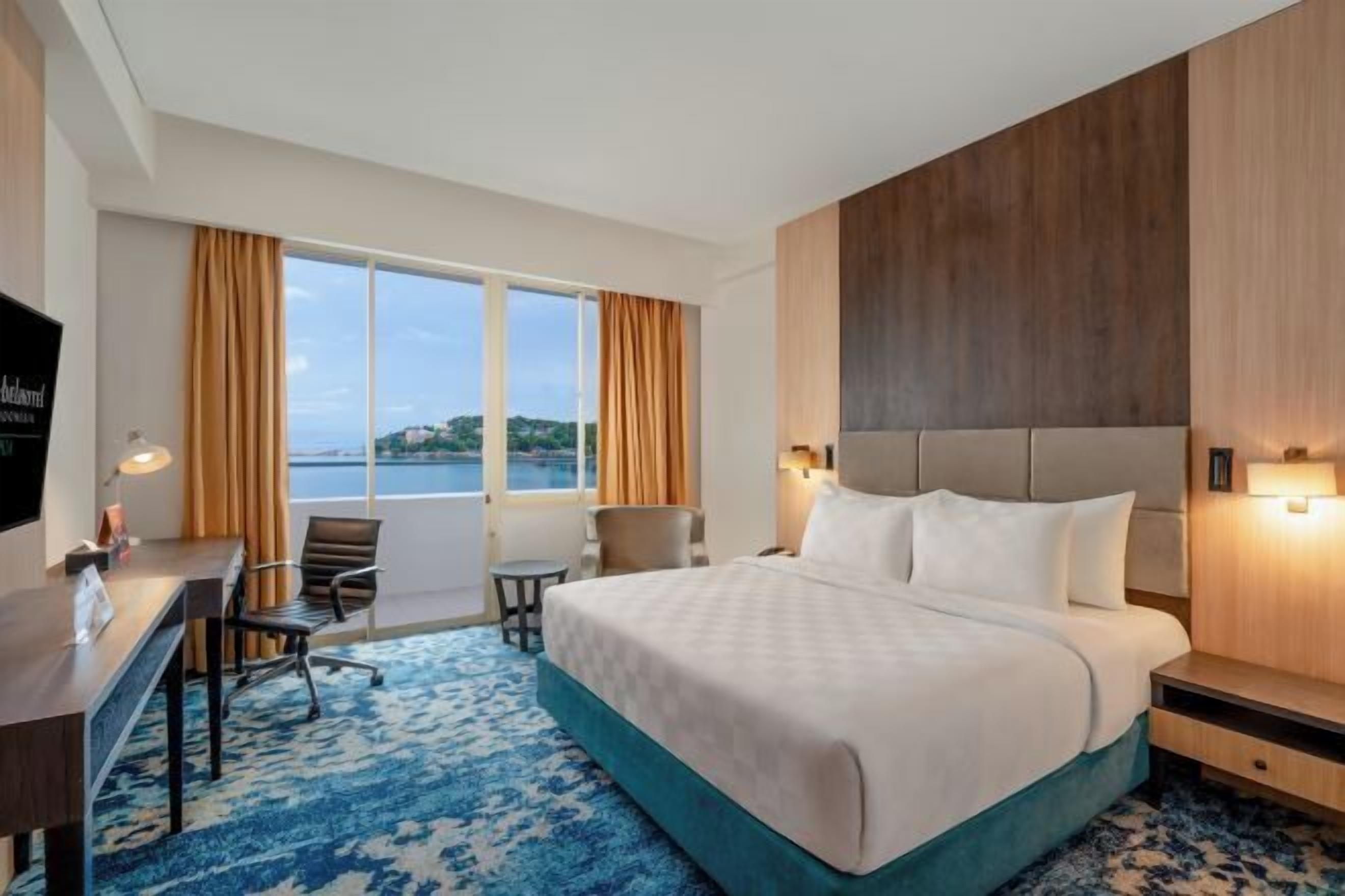 Habitación de hotel con cama, escritorio, silla y un gran ventanal con cortinas.
