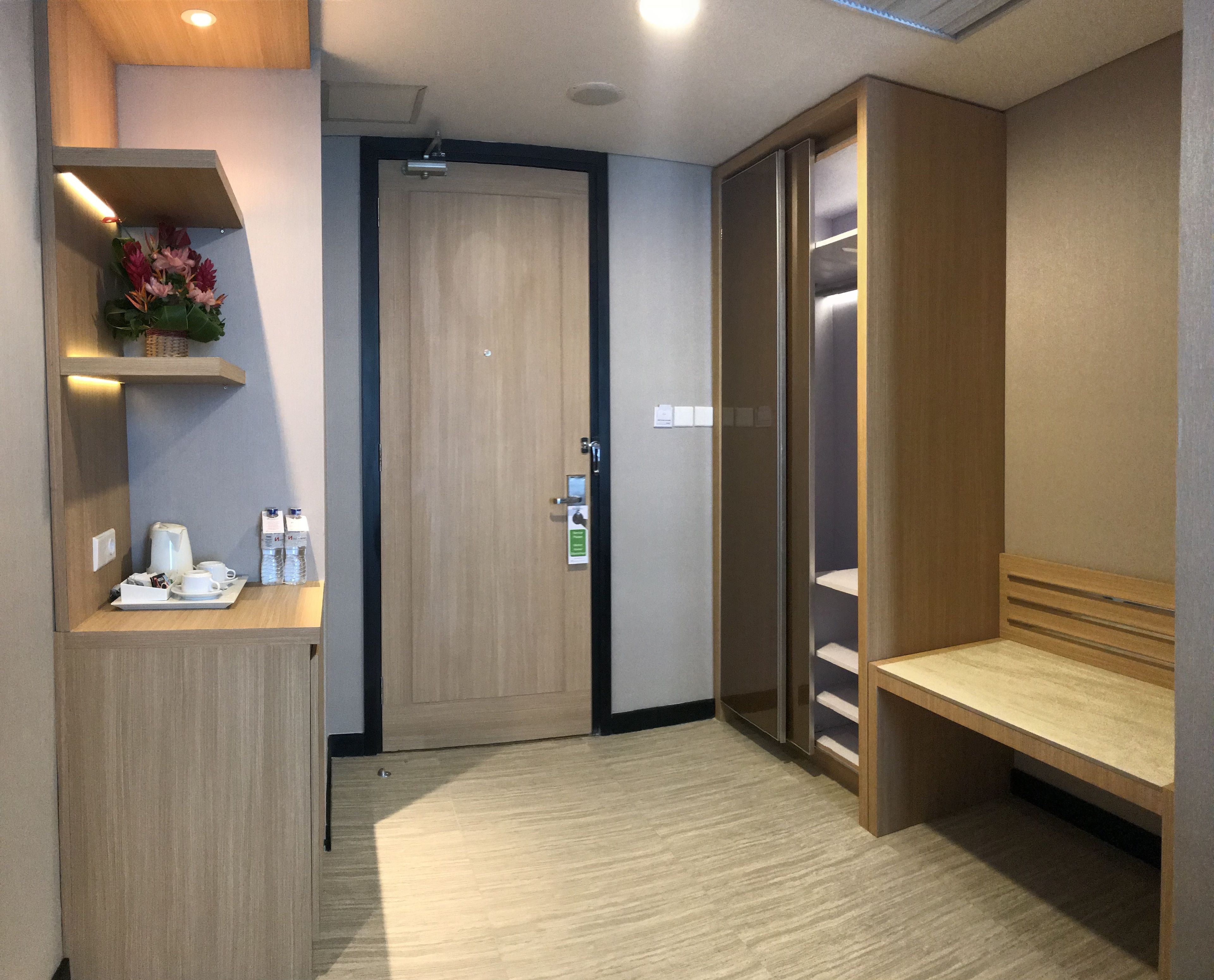 A hotel room with a wooden door, a small table with a coffee maker and water bottles, a shelf with flowers, and a wooden bench.